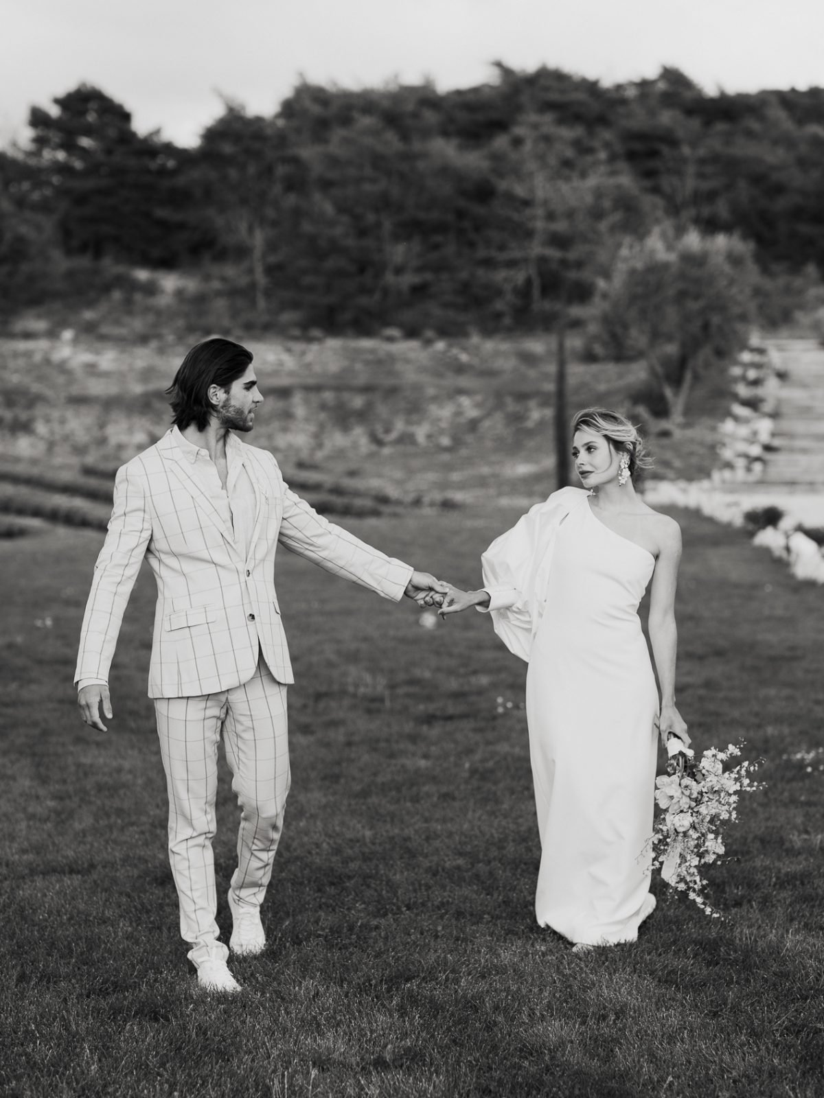 Portrait romantique d'un couple de mariés qui marchent en se tenant la main. Ils se regardent amoureusement. Mariage d'été, photographie de mariage en Provence, France, élégante et intemporelle, en noir et blanc, réalisée par Charlotte Marette