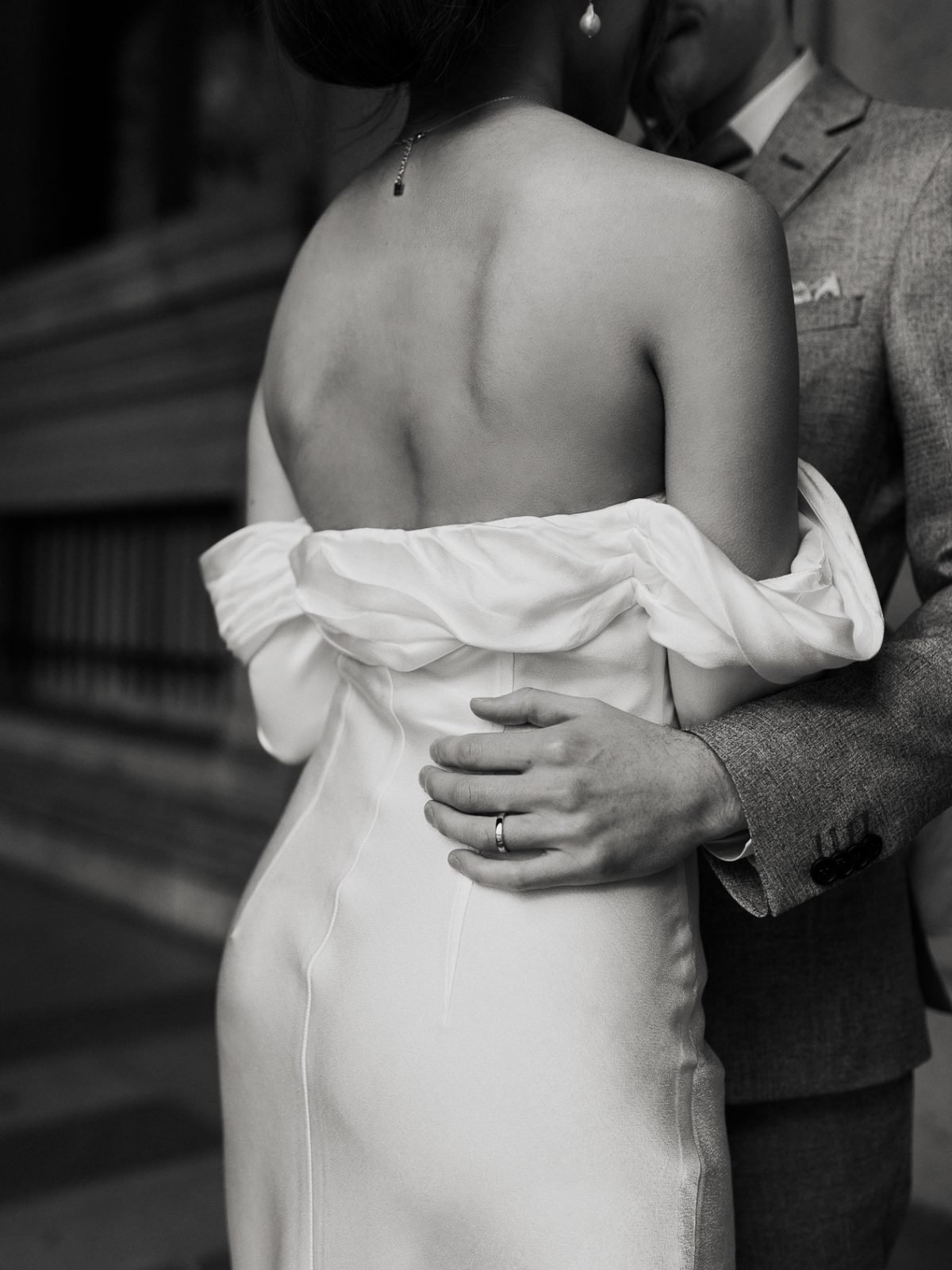 Couple marié s'enlaçant dans les galeries du Louvre à Paris. La femme est de dos, la main de son mari est posée sur sa taille, on voit l'alliance en or blanc. Photographie de mariage romantique, élégante et intemporelle en noir et blanc. Photographie réalisée par Charlotte Marette